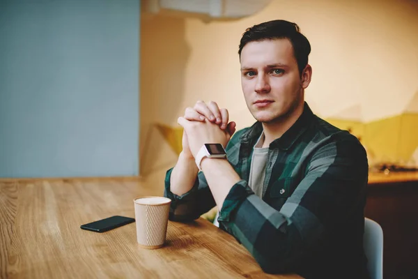 Portrait of sad young male looking at camera feeling alone and need conversation about problem,worried hipster guy thinking about solutions sitting at table with coffee cup in cafeteria on leisure