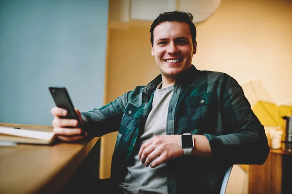 Portrait of cheerful teen male holding cellular satisfied with wireless connection for chatting and sharing content leisure,happy hipster guy looking at camera using telephone for blogging and networking