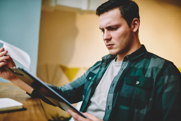 Serious young male coworker disappointed with accountings in report holding documents planner, professional designer reading and analyzing information for project upset with sketches in office