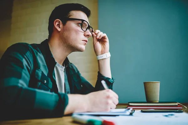 Smart young hipster guy thinking about problem solution doing homework in coworking space,pensive male student in eyewear concentrated on creative idea for coursework making notes and planning