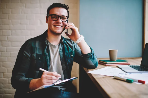 Portrait of cheerful man satisfied with telephone consultancy while noting information for studying,successful hipster guy making telephone call during writing article on remote job in coworking space