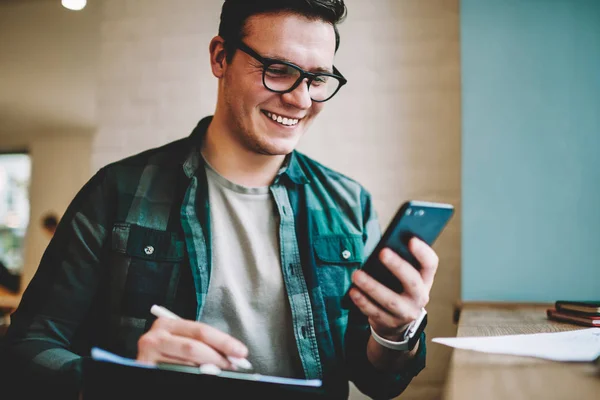 Happy male in spectacles satisfied with getting discount for 4G connection usage chekcing mail on mobile phone, cheerful hipster guy confirming new friend in social network using smartphone