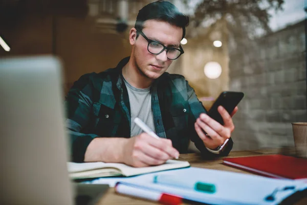 Pensive male student in eyewear writing information in notebook holding telephone, concentrated man freelancer noting contact from mobile telephone sitting in coworking office with technology