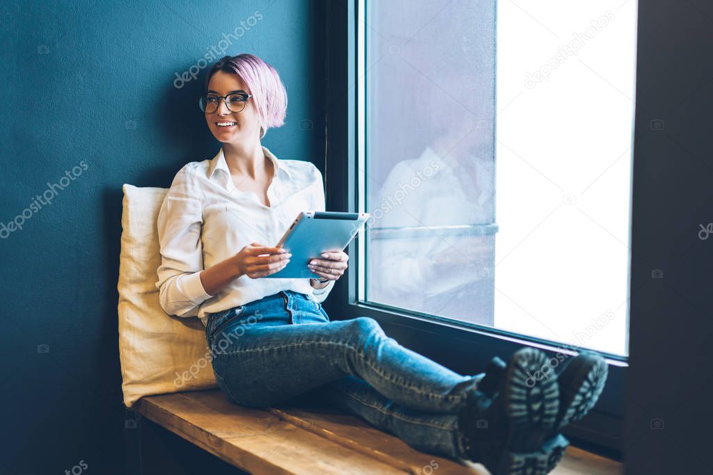 Beautiful successful talented female student dressed in trendy outfit laughing while spending leisure time indoors.Cheerful hipster girl having fun during using modern device sitting next to window