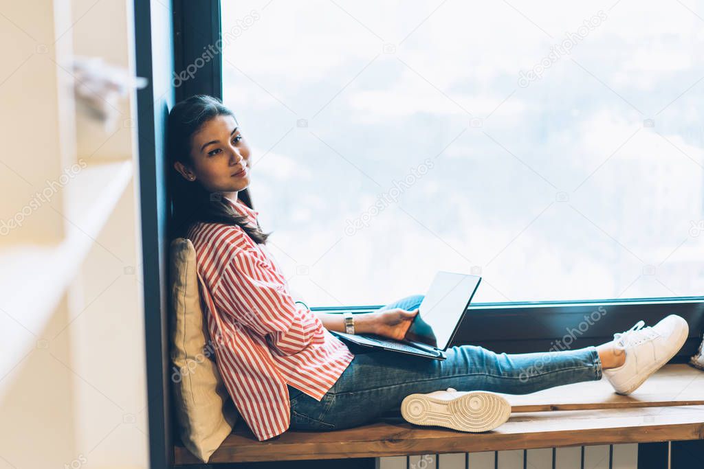 Young charming happy hipster girl dressed in trendy outfit dreaming while recreating sitting next to window with copy space.Cute positive female blogger waiting for downloading files from network