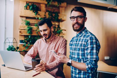 Neşeli erkek bu kameraya bakarak ve arkadaşı dizüstü bilgisayar, müreffeh çalışan olarak meslektaşım ile işbirliği Web'de bilgi okuma sırasında telefon tutarak geliştirici