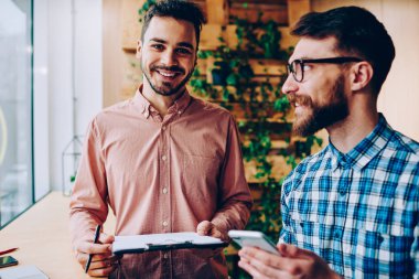 Klasör başarılı başlangıç eğitim hakkında arkadaşıyla danışmanlık sırasında kameraya bakarak hipster adam gülümseyen meslektaşı ile işbirliği için planları ile tutarak yakışıklı neşeli adam portresi