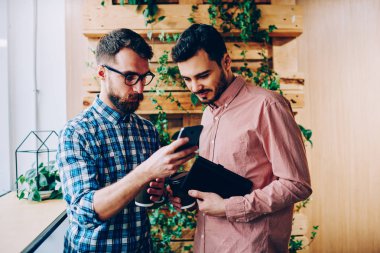 Genç erkek arkadaşlar akıllı telefonlar için yeni app kontrol yüklemek, hipster adam danışmanlık meslektaşım rezervasyon hakkında onun cep üzerinde bilgi gösteren Web sitesinde için dosyaları indirmek