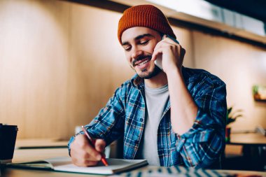 Neşeli erkek yaratıcı tasarımcı çizim kroki telefon konuşmayı, üniversite librar otururken fikirleri tartışmak için arkadaşıma telefon görüşmesi yapma hipster adam gülümsüyor not defterinde