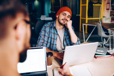 Düşünceli hipster adam erkek görüntü kırpılmış üniversitede geliştirici hakkında bir şey colleagu ile çalışma sürecinde hayal öğrenme için dizüstü bilgisayar başında otururken kameraya bakıyor