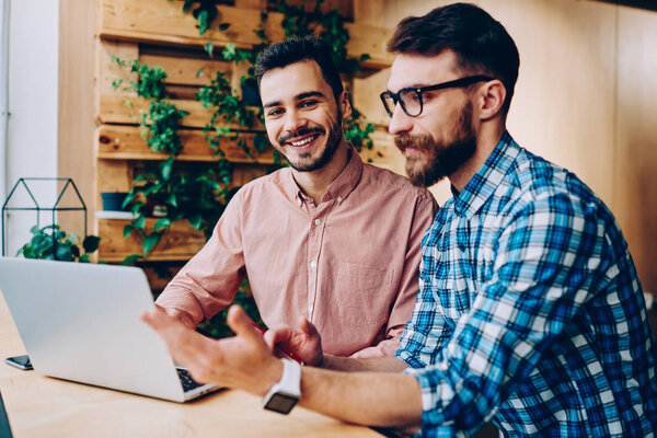 Portrait of cheerful handsome man looking at camera while his friend explaining information and idea  while cooperating, skilled male programmers working together on project using laptop computer 