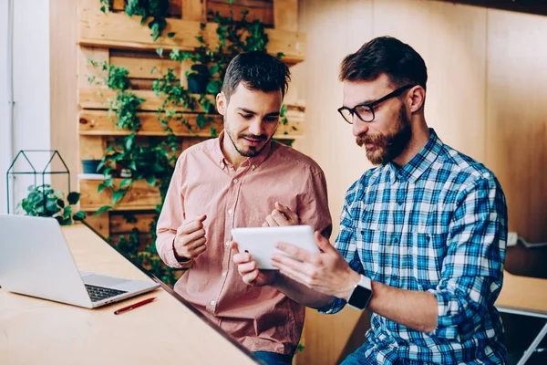 Young hipster guys watching tutorial on digital tablet preparing for exams together using technology,men browsing information for project on touchpad cooperating and talking in coworking office