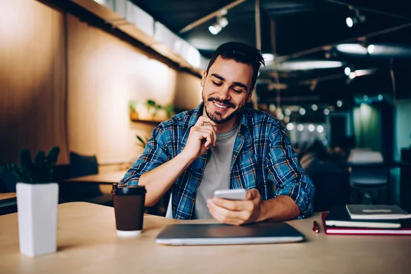 Smiling hipster guy reading new message in social networks confirming new friend on smartphone, positive young man satisfied with banking app on mobile phone checking balance while learnin