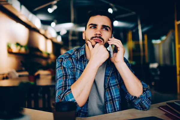 Contemplative hipster guy thinking aver plans for meeting while talking on telephone with friend in cafe, pensive young man waiting for connection with customer support line on mobile phon