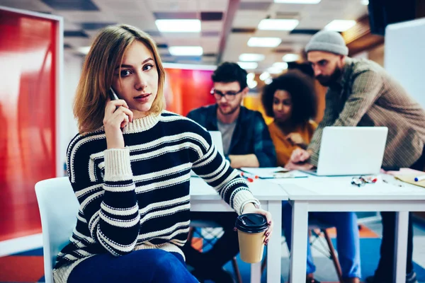 Portrait of attractive student with short haircut talking on smartphone while looking at camera.Blonde hipster girl communicating with operator on modern smartphone sitting in office interior