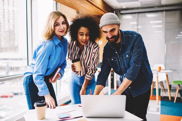Half length portrait of attractive positive blonde standing next to smart colleagues during collaboration in office.Hipster girl looking at camera while partners searching useful information on laptop