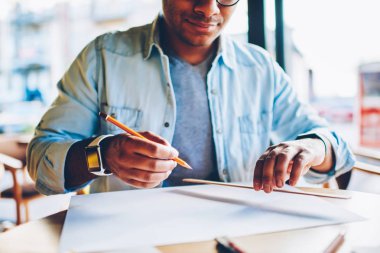 Kırpılmış görüntü akıllı yaratıcı erkek mimar planları üzerinde kapalı oturma sırasında çalışma trendy kot elbise giymiş. Kalem ve kural skeçler oluşturulurken holding adam eller odaklanmak