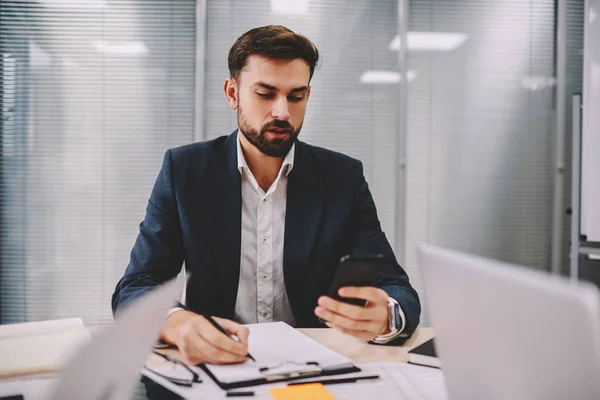 Pensive proud ceo rewriting information data from smartphone device working in office interior.Bearded businessman reading incoming notification on telephone while making accounting reports