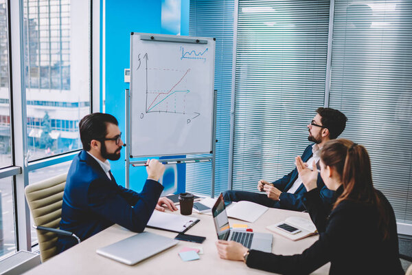 Prosperous proud ceo discussing financial profit drawn in graphic on flipchart collaborating with professional employees.Businessman with experts working on strategy at meeting table in office