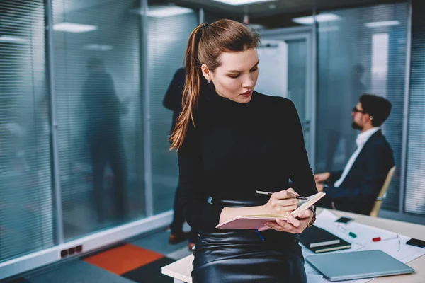 Young charming contemplative female graphic designer dressed in formal wear writing down creative ideas to notepad while sitting on table.Intelligent woman employee making notes during work indoors