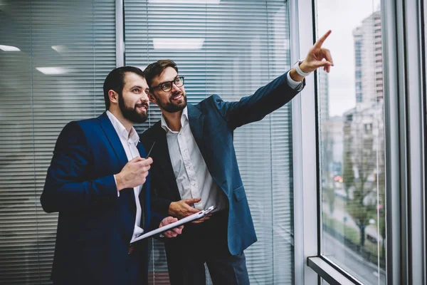 Handsome successful male entrepreneurs dressed in formal clothes having fun and looking outside during informal meeting indoors.Young bearded man pointing outside for showing to friend funny thing