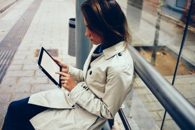 Parmak sahte uygulama açık havada tramvay durağı üzerinde oturan 4 g internet bağlantısı üzerinden yüklemelerde seçme modern Dokunmatik ped cihaz ekranı ile dokunmadan dalgın kadın hipster blogger yan görünüm