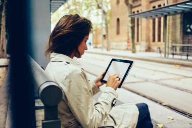 Genç kadın sahte alanı Web sitesi içerik tramvay durağı üzerinde oturan için modern Dokunmatik ped cihaz üzerindeki metin yazarak. Hipster blogger tablet ile boş perde reklam için web sayfasında bir haber okuma