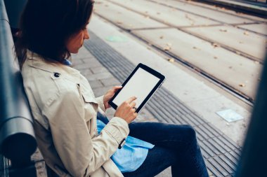Şık giyimli genç kadın sahte ekranı modern dokunmatik yüzey üzerinde parmak ile dokunmadan yan görünüm bağlı 4 g için taşıma tramvay durağı üzerinde oturup beklerken online alışveriş yaparken Internet
