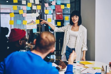 Atölye çok kültürlü grup öğrenci için iletken ve daha iyi öğrenmek için yabancı kelimelerle renkli çıkartmaları ile cam duvara işaret ederken kameraya gülümseyen genç kadın portresi