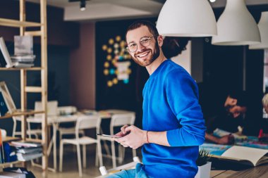 Portrait of cheerful young man in casual wear smiling at camera while updating software on modern touch pad device using wireless 4G internet.Positive hipster guy installing app on tablet in office clipart
