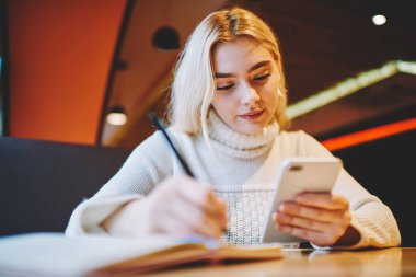 Young woman making notes using organizing application on cellular for planning tasks and learning process, female writing in notepad information from web page browsed on smartphone in cafe interior clipart