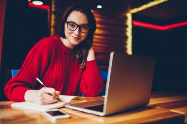 Gülümseyen genç kız öğrenci ödev görev kafe iç dizüstü bilgisayar ile olumlu yaratıcı kadın blogger yazma makale içinde not etmek için sosyal ağ yayın yapıyor portresi