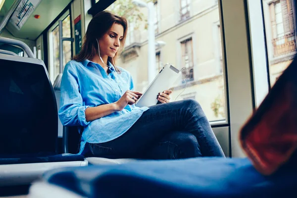 Female blogger listening music player online on website in earphones connected to digital tablet sitting in tram.Hipster girl watching video in networks on touch pad using public wireless internet