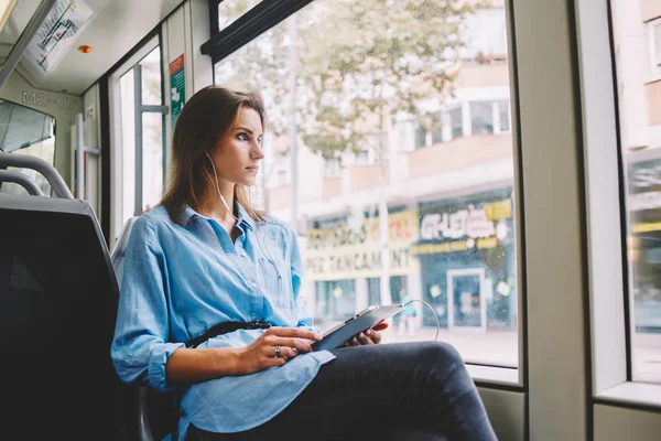 Pondering young woman looking out of window and listening lyric music on radio website in modern earphones connected to tablet device sitting in tram and using public wireless internet connection