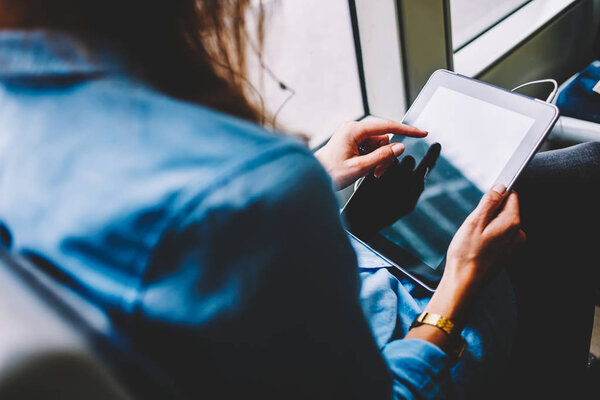 Cropped back view of young woman holding modern tablet in hands and touching with finger on display using free transport wifi in tram.Female passenger in earphpnes choosing music app on touch pad