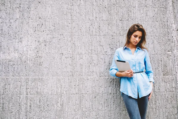 Young woman dressed in casual blue shirt looking down while holding modern tablet in hands.Hipster girl with digital touch pad device standing outdoors near concrete wall with publicity area