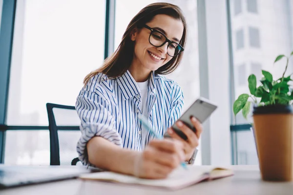 千年の少女眼鏡 宿題を書きながらスマート フォンで収入メッセージを読んで Blo の記事出版物の創造的なアイデアのメモを作ってメールをチェック女性従業員の笑顔 — ストック写真