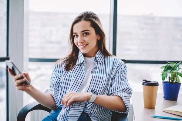 Portrait of smiling pretty employee sitting at desktop holding mobile phone for chatting on break, cheerful casually dressed woman looking at camera using smartphone and wireless internet in office