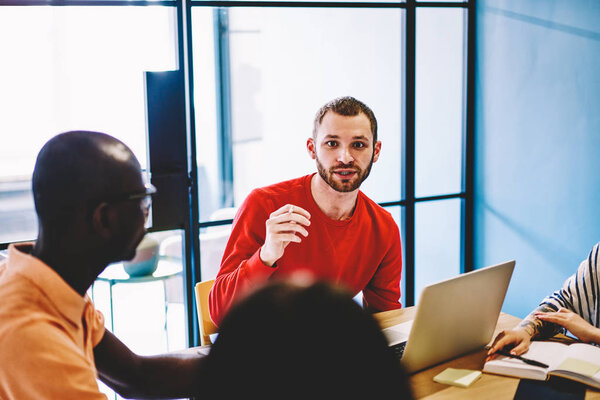 Multicultural group of young people dressed in smart casual wear teamworking on common project in office.Portrait of male leader looking at camera while sitting at laptop computer during briefing