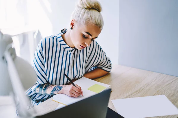 Pensive blonde caucasian young woman in stylish casual wear writing down plan organization in notepad sitting at desktop in office.Creative female designer making notes of publication in notebook