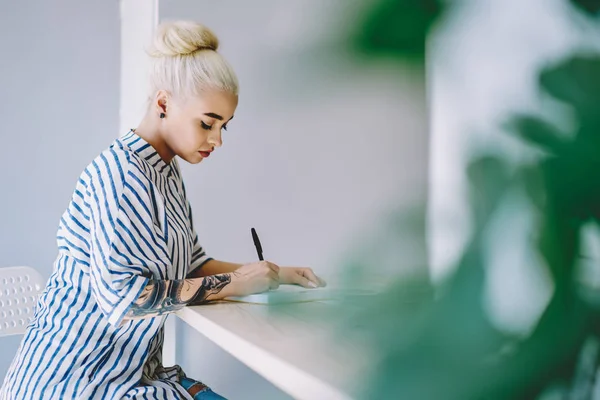 Pensive blonde young woman dressed in casual wear writing down organization plan of working process in notepad sitting at desktop in modern office.Smart student doing homework in coworking space