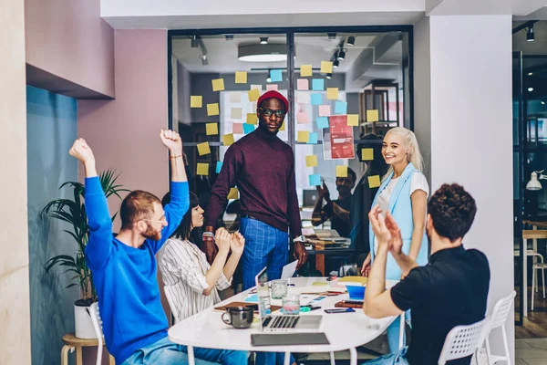 Portrait of multicultural hipster students dressed in smart casual wear looking at camera while celebrating victory and completing project sitting at meeting table in modern office interior