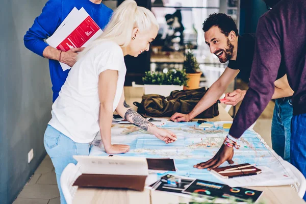 Positive hipster guys in smart casual wear laughing while teamworking and marking on geography map choosing countries during brainstorming.Cheerful young people laughing standing at desktop with atlas