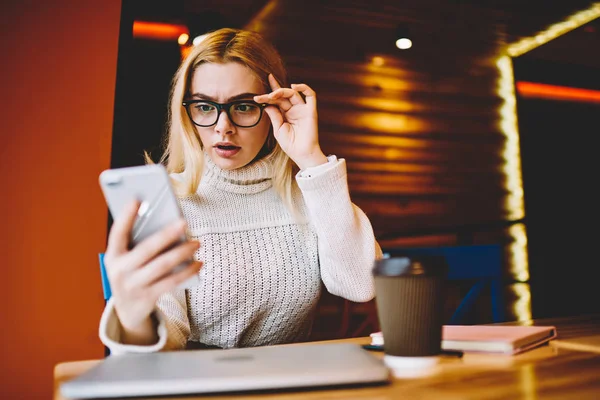 Shocked nervous woman in eyewear can\'t believe in bad information getting on email holding smartphone,stressed hipster girl disappointed with news feeling irritated reading message on telephon
