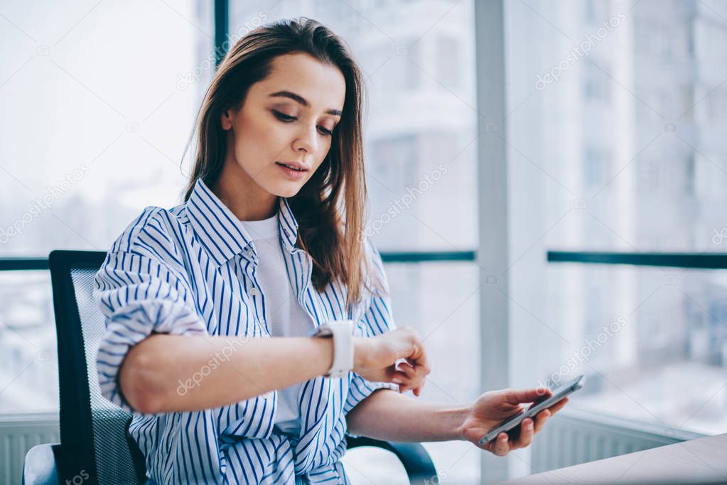 Young attractive female checking time of wrist watches while sitting in office interior holding mobile phone in hands. Brunette woman employee waiting for coffee break during long work day