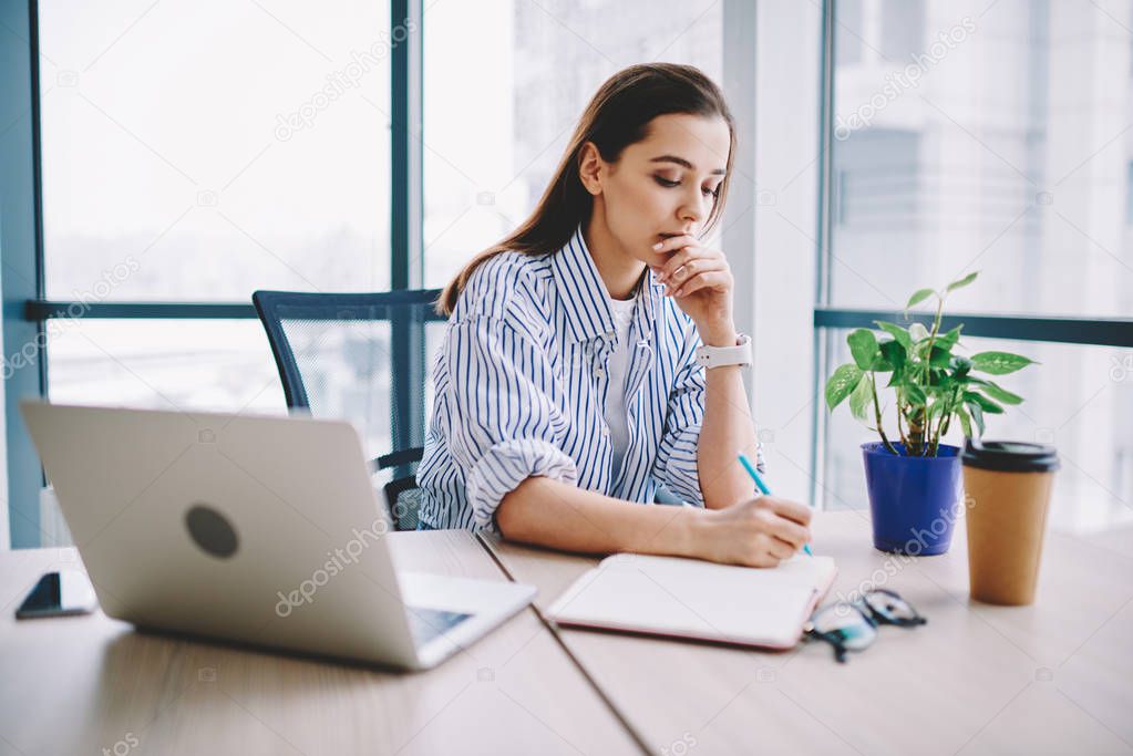 Concentrated on work woman making notes of planning project strategy sitting at desktop with netbook,creative pensive female web designer writing ideas in notebook organizing process in offic