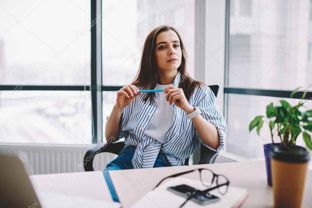 Serious brunette young woman sitting at desktop looking away pondering on idea for working project, creative female designer puzzled on business spending time at loft interior office holding pe