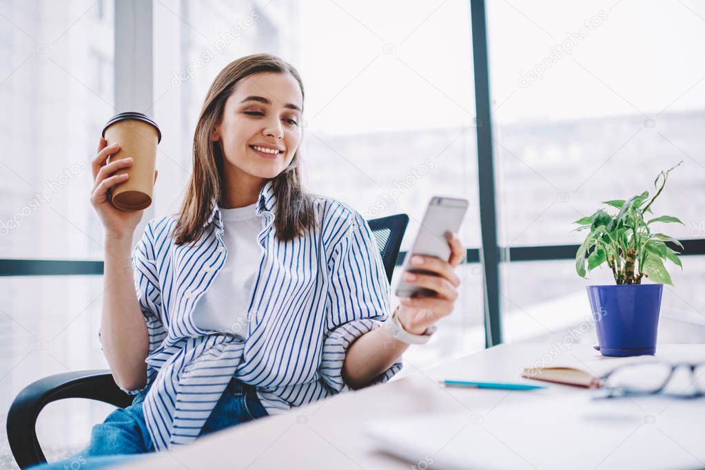 Cheerful young woman watching video in social networks on smartphone during break holding coffee cup, smiling  hipster girl sending text message via mobile phone sitting at desktop in offic