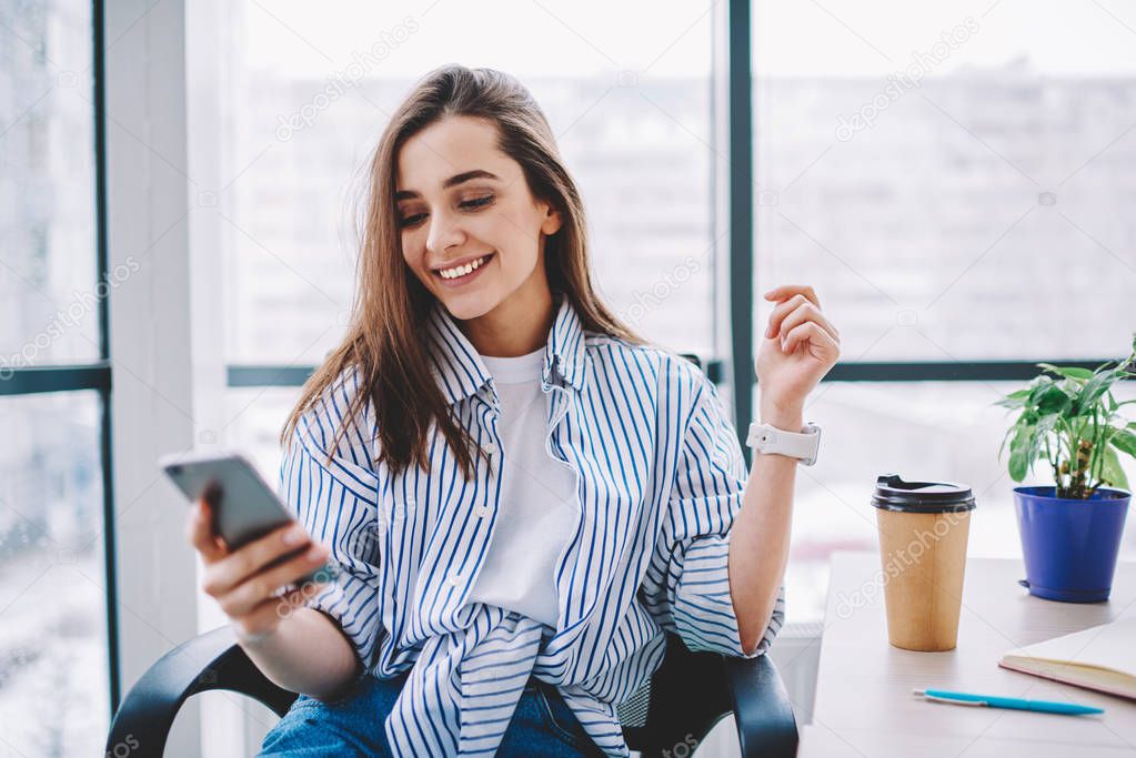 Cheerful young woman checking new feed and content in social networks via smartphone during coffee break in office,happy cute female blogger satisfied with followers messages sitting at deskto