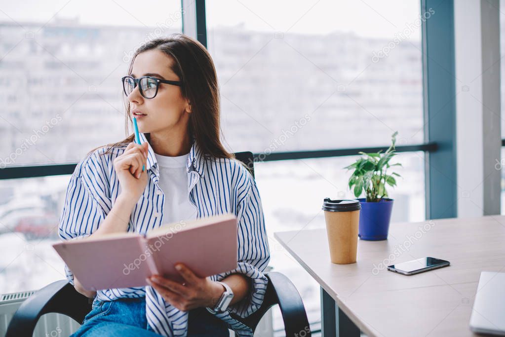 Pensive female author in eyewear looking away concentrated on idea for article sitting at desktop,skilled professional woman journalist pondering on publication holding pen and notebook in office